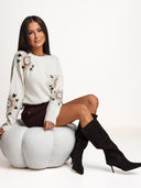 Sweter ecru OH HONEY WINTER ROSE z wełną merino i kaszmirem na siedzącej modelce — kwiatowy wzór na przodzie i rękawach.